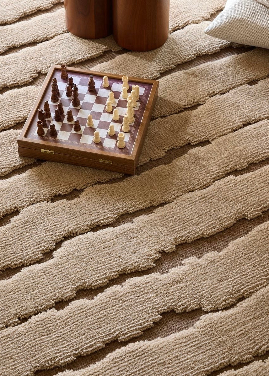Small wooden chessboard with pieces on a textured beige carpet with a wooden side table and pillow.